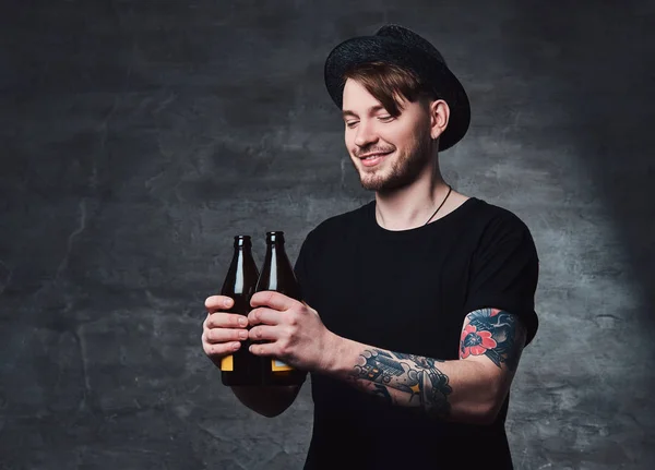 Portrait of an attractive bearded male with tattooed arms, dressed in a black t-shirt and hat holds bottled beer.