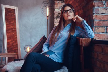 Gözlük ve mavi gömlek tutar tablet bir esmer kız sevimli ve çatı tasarımı ile bir odada bir kanepede oturmuş portre.