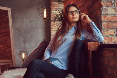 Gözlük ve mavi gömlek tutar tablet bir esmer kız sevimli ve çatı tasarımı ile bir odada bir kanepede oturmuş portre.