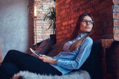 Gözlük ve mavi gömlek tutar tablet bir esmer kız sevimli ve çatı tasarımı ile bir odada bir kanepede oturmuş portre.