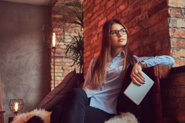 Esmer işkadını tablet bir gözlük ve mavi gömlek tutar olarak çekici ve çatı tasarımı ile bir odada bir kanepede oturmuş portre.