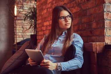 Esmer işkadını tablet bir gözlük ve mavi gömlek tutar olarak çekici ve çatı tasarımı ile bir odada bir kanepede oturmuş portre.