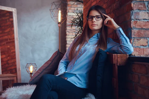 眼鏡と青シャツが保持しているタブレットのブルネットの女の子を魅力的なとロフト デザイン ルームでソファの上に座っての肖像画 — ストック写真
