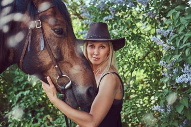 Siyah elbise ve bir at Çiçek bahçesinde sarılma şapka giyen sevimli güzel sarışın.