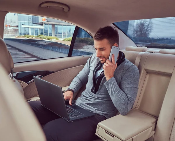 Красивый Бизнесмен Элегантной Одежде Работающий Ноутбуке Разговаривающий Телефону Сидя Машине — стоковое фото