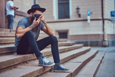 Yakışıklı hippi erkek bir şık sakal ve kollarını dövme ile rahat kıyafetler giymiş ve şapka bir kamera tutan, merdiven üstünde oturuyor.