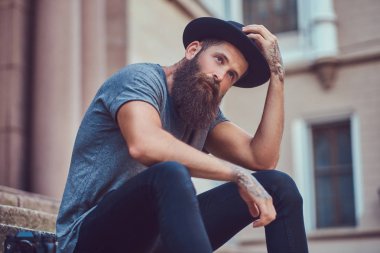 Kollarını bir dövme ile şık sakallı yakışıklı hippi erkek rahat kıyafetler giymiş ve şapka merdivenlerinde oturur.