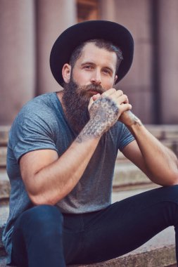Kollarını bir dövme ile şık sakallı yakışıklı hippi erkek rahat kıyafetler giymiş ve şapka merdivenlerinde oturur.