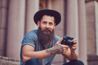 Yakışıklı hippi erkek bir şık sakal ve kollarını dövme ile rahat kıyafetler giymiş ve şapka bir kamera tutan, merdiven üstünde oturuyor.