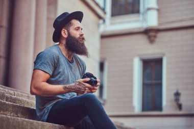 Yakışıklı hippi erkek bir şık sakal ve kollarını dövme ile rahat kıyafetler giymiş ve şapka bir kamera tutan, merdiven üstünde oturuyor.