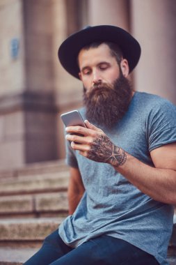 Yakışıklı hipster erkek şık sakal ve kolunda bir dövme ile rahat kıyafetler ve merdiven üstünde otururken smartphone kullanarak şapka giymiş.