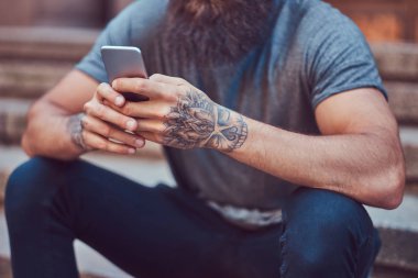 Yakışıklı hipster erkek şık sakal ve kollarını bir dövme ile rahat kıyafetler ve merdiven üstünde otururken smartphone kullanarak şapka giymiş.