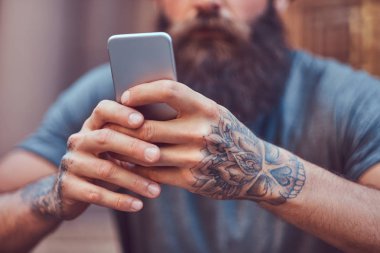 Yakışıklı hipster erkek şık sakal ve kollarını bir dövme ile rahat kıyafetler ve merdiven üstünde otururken smartphone kullanarak şapka giymiş.
