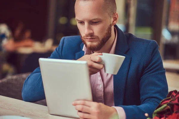 Kafeteryanın dışında tablet bilgisayarda çalışan yakışıklı Kızıl saçlı işadamı bir şık saç ve sakal bir mavi ceket ve pembe gömlek giymiş.