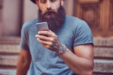 Yakışıklı hipster erkek şık sakal ve kollarını bir dövme ile rahat kıyafetler ve merdiven üstünde otururken smartphone kullanarak şapka giymiş.
