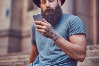 Yakışıklı hipster erkek şık sakal ve kollarını bir dövme ile rahat kıyafetler ve merdiven üstünde otururken smartphone kullanarak şapka giymiş.