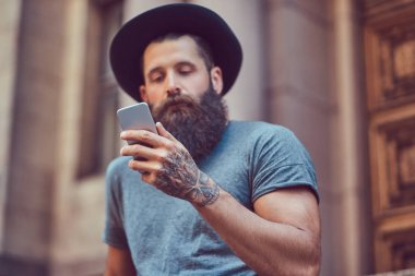 Yakışıklı hipster erkek şık sakal ve kollarını bir dövme ile rahat kıyafetler ve merdiven üstünde otururken smartphone kullanarak şapka giymiş.