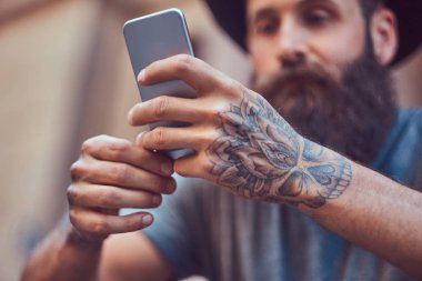 Yakışıklı hipster erkek şık sakal ve kollarını bir dövme ile rahat kıyafetler ve merdiven üstünde otururken smartphone kullanarak şapka giymiş.