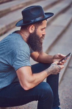 Yakışıklı hipster erkek şık sakal ve kollarını bir dövme ile rahat kıyafetler ve merdiven üstünde otururken smartphone kullanarak şapka giymiş.