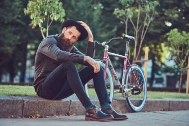 Yakışıklı hipster seyahat bir şık sakal ve kollarını bir Bisiklete binmek sonra dinlenme kaldırımda oturan bir çanta, şapka ve rahat kıyafetler giymiş dövme.