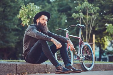 Yakışıklı hipster seyahat bir şık sakal ve kollarını bir Bisiklete binmek sonra dinlenme kaldırımda oturan bir çanta, şapka ve rahat kıyafetler giymiş dövme.