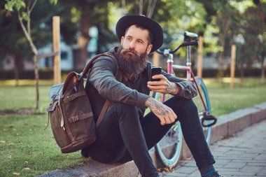 Bisiklete binmek sonra dinlenme kaldırımda oturan smartphone kullanarak yakışıklı hipster seyahat bir şık sakal ve kollarını bir çanta, şapka ve rahat kıyafetler giymiş dövme.
