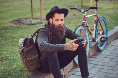Bisiklete binmek sonra dinlenme kaldırımda oturan smartphone kullanarak yakışıklı hipster seyahat bir şık sakal ve kollarını bir çanta, şapka ve rahat kıyafetler giymiş dövme.