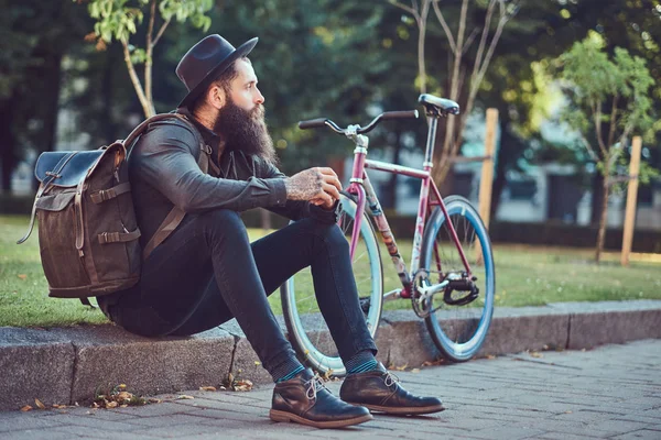 カジュアルな服と自転車に乗って後休んで 歩道の上に座って バッグと帽子に身を包んだスタイリッシュなひげと彼の腕のタトゥーとハンサムな流行に敏感な旅行者 — ストック写真