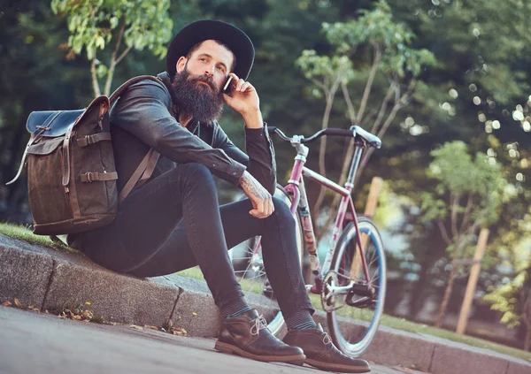 英俊的时髦的旅行者与一个时髦的胡子和纹身在他的胳膊穿着休闲衣服和帽子与袋子 使用智能手机 坐在人行道上 休息后骑自行车 — 图库照片
