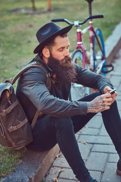 自転車に乗って後休んで 歩道の上に座って スマート フォンを使用してスタイリッシュなひげとカジュアルな服やバッグ 帽子に身を包んだ彼の腕のタトゥーとハンサムな流行に敏感な旅行者 — ストック写真