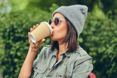 Hipster kadın bir şehir parkta bir bankta oturmuş kahve ile şapkalı. Doğada rahatlatıcı.