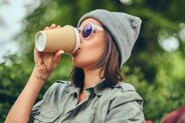 Hipster kadın bir şehir parkta bir bankta oturmuş kahve ile şapkalı. Doğada rahatlatıcı.