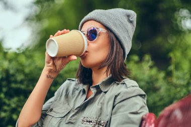 Hipster kadın bir şehir parkta bir bankta oturmuş kahve ile şapkalı. Doğada rahatlatıcı.