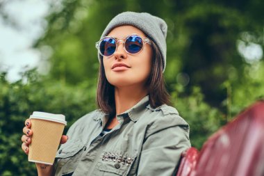 Hipster kadın bir şehir parkta bir bankta oturmuş kahve ile şapkalı. Doğada rahatlatıcı.