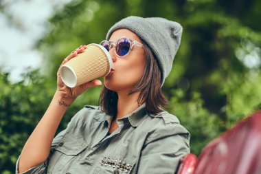 Hipster kadın bir şehir parkta bir bankta oturmuş kahve ile şapkalı. Doğada rahatlatıcı.