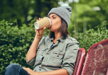 Hipster kadın bir şehir parkta bir bankta oturmuş kahve ile şapkalı. Doğada rahatlatıcı.