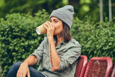 Hipster kadın bir şehir parkta bir bankta oturmuş kahve ile şapkalı. Doğada rahatlatıcı.