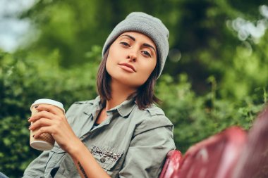 Hipster kadın bir şehir parkta bir bankta oturmuş kahve ile şapkalı. Doğada rahatlatıcı.