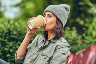Hipster kadın bir şehir parkta bir bankta oturmuş kahve ile şapkalı. Doğada rahatlatıcı.