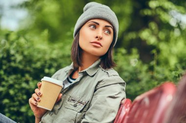 Hipster kadın bir şehir parkta bir bankta oturmuş kahve ile şapkalı. Doğada rahatlatıcı.