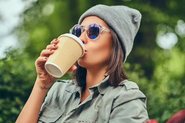 Hipster kadın bir şehir parkta bir bankta oturmuş kahve ile şapkalı. Doğada rahatlatıcı.