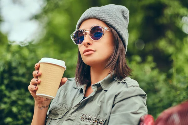 Hipster kadın bir şehir parkta bir bankta oturmuş kahve ile şapkalı. Doğada rahatlatıcı.