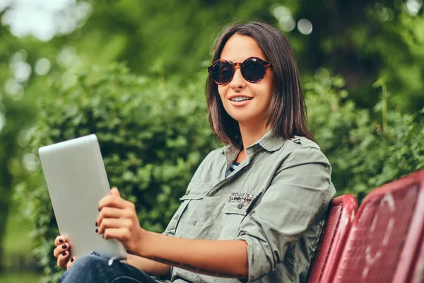 Hipster kadın bir şehir parkta bir bankta oturmuş tablet ile. Doğada rahatlatıcı.