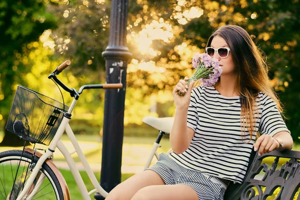 Retrato de uma morena atraente se senta em um banco com bicicleta — Fotografia de Stock