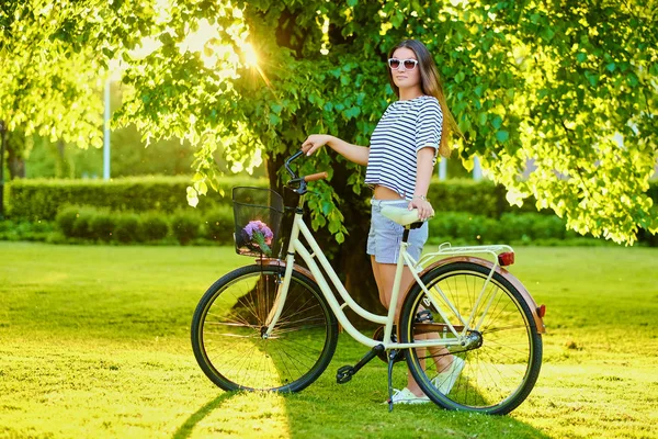 Hermosa morena se para en el césped verde con bicicleta en el parque — Foto de Stock