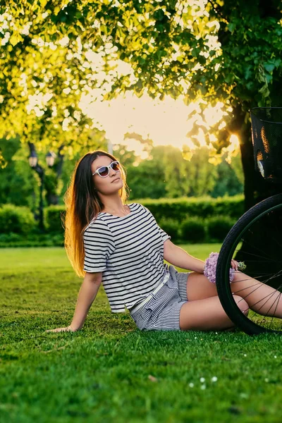 Hermosa morena se sienta en el césped verde con bicicleta y sostiene — Foto de Stock