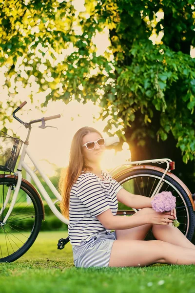 Schöne Brünette sitzt mit Fahrrad auf dem grünen Rasen und hält — Stockfoto