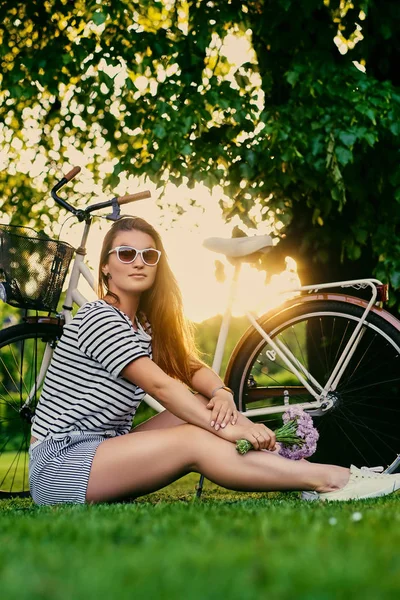 Schöne Brünette sitzt mit Fahrrad auf dem grünen Rasen und hält — Stockfoto