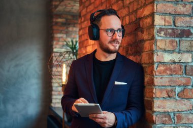 Bir tuğla duvara tablet ile müzik dinleme şık takım elbise giyen bir yakışıklı sakallı erkek portresi.