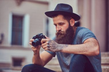 Yakışıklı hippi erkek bir şık sakal ve kollarını dövme ile rahat kıyafetler giymiş ve şapka bir kamera tutan, merdiven üstünde oturuyor.